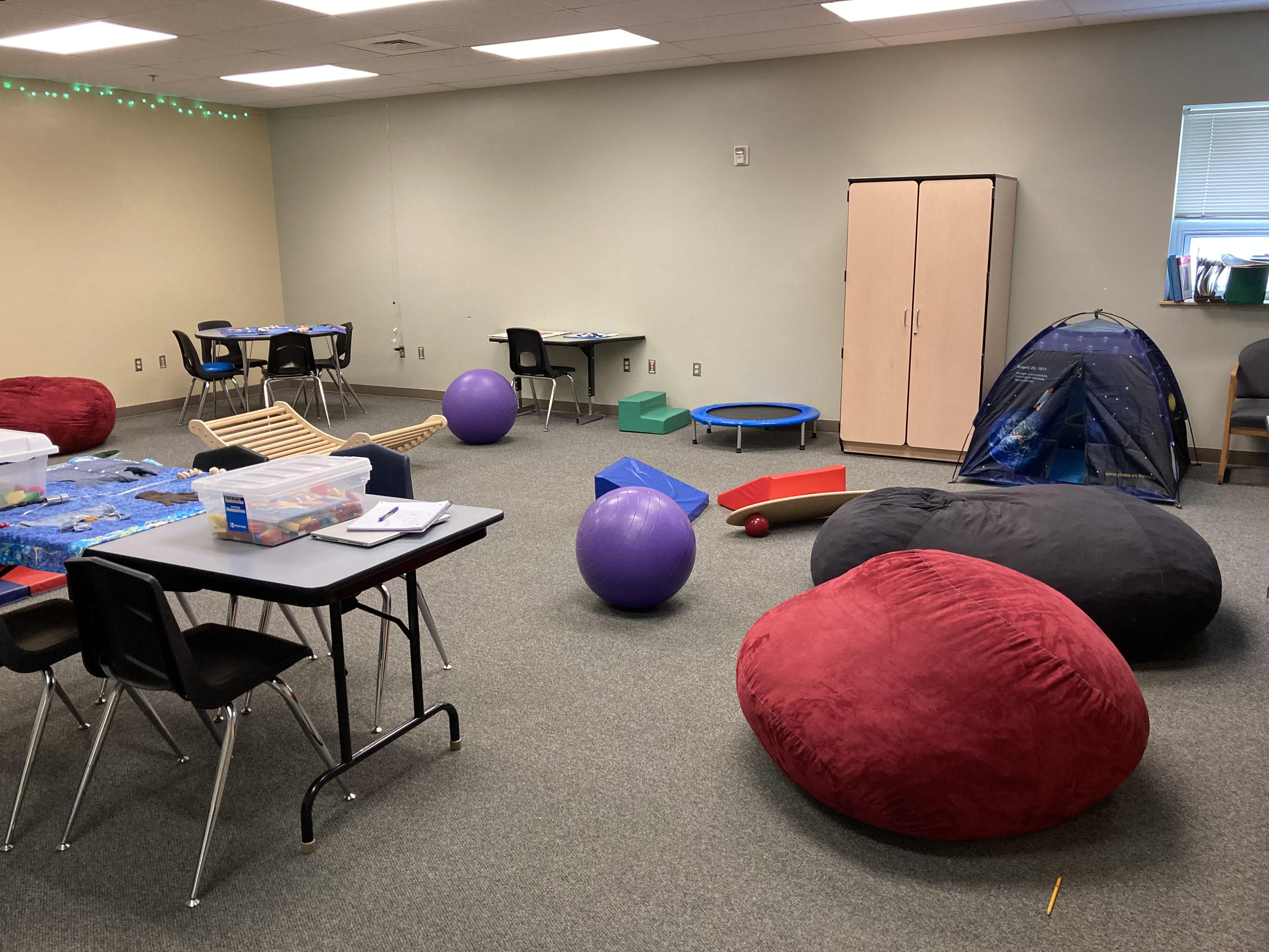 A classroom featuring tables filled with sensory items, exercise balls, a tent, bean bag chairs